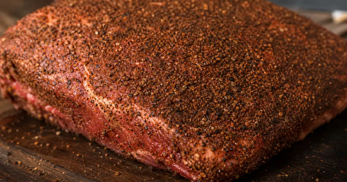 A raw beef brisket generously coated in a coarse dark BBQ rub, resting on a rustic wooden cutting board