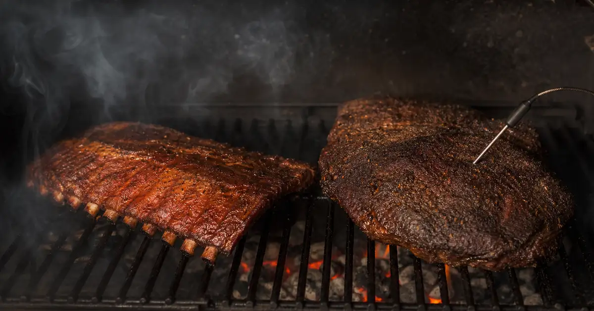 Ribs and a brisket resting on cast-iron grill grates with drifting wood smoke and a temperature probe