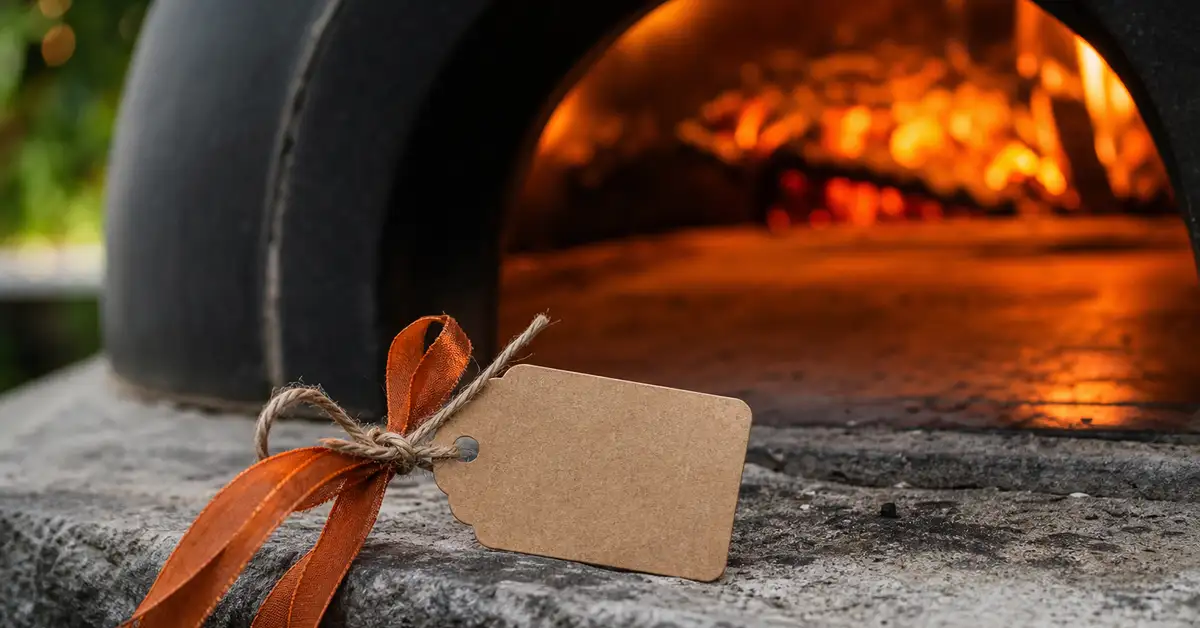 A small kraft-paper gift tag tied with ember-orange ribbon resting beside the glowing mouth of a portable outdoor pizza oven