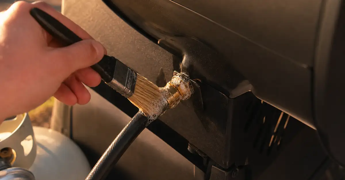 Close-up of hands performing a soapy-water leak check on a propane grill hose connection, warm afternoon light