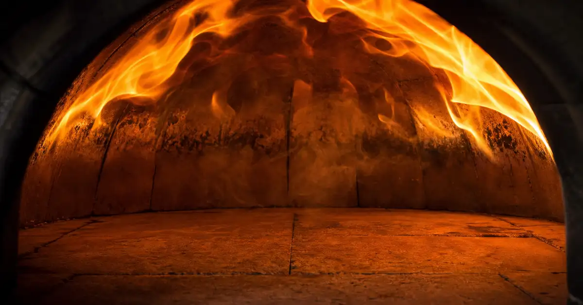 Close-up of orange flame rolling across the curved dome ceiling inside a lit pizza oven above an empty stone floor