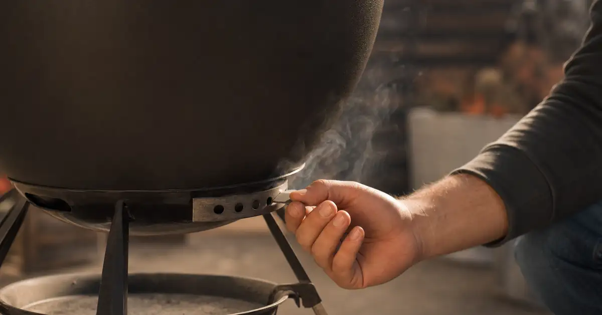 A hand adjusting the bottom vent damper on a charcoal kettle grill to control airflow and temperature