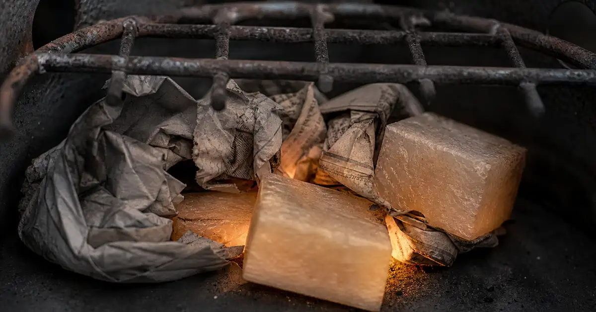 Macro close-up of crumpled newspaper and paraffin fire-starter cubes tucked beneath the grate of an empty charcoal chimney starter