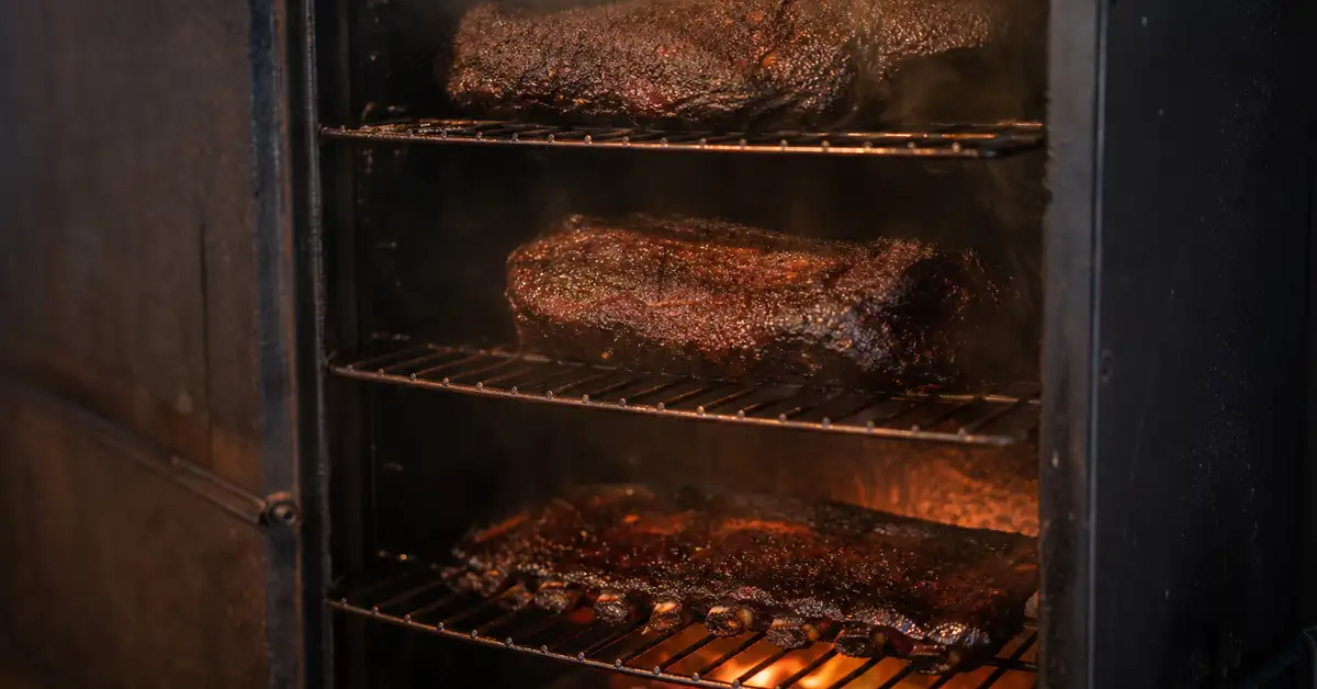 Multiple racks stacked inside an open vertical smoker cabinet with warm smoke curling between them