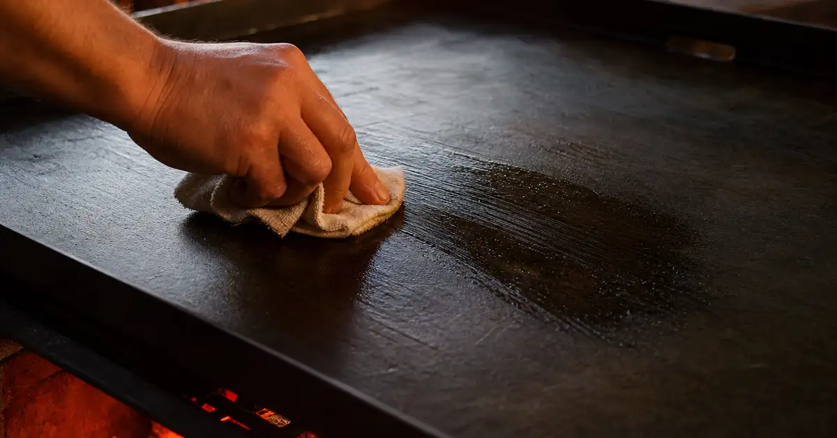 Wiping a thin layer of oil across a flat steel griddle surface with a folded cloth