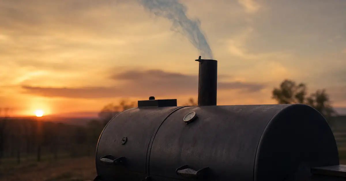 Thin blue-gray smoke drifting from the smokestack of a dark offset smoker at golden hour